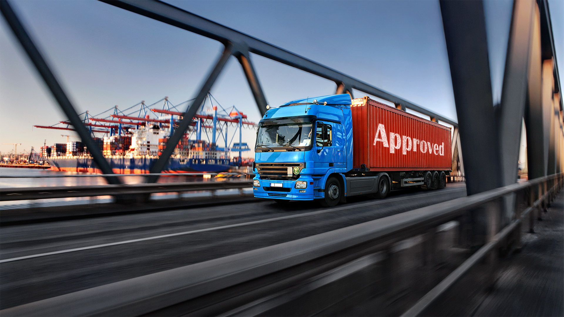 Ein blau-roter LKW mit einem seitlichen "Approved"-Aufdruck fährt über eine Brücke, im Hintergrund Hafenanlagen